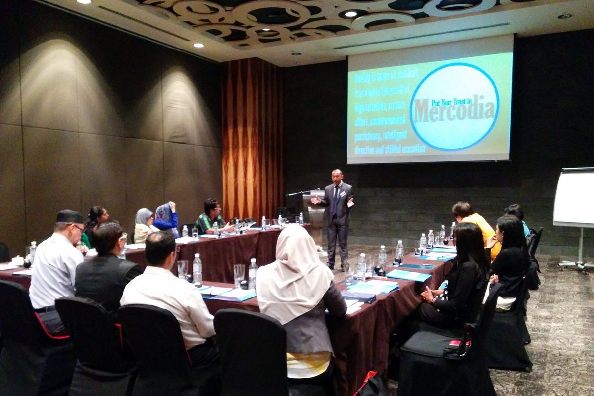 A speaker presenting to participants seated at long tables in a hotel meeting room, with a Mercodia slide on the screen