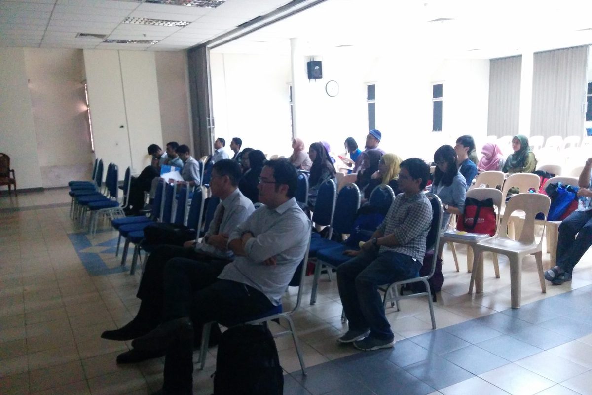 Participants seated in rows of blue and beige chairs in a bright hall during a session
