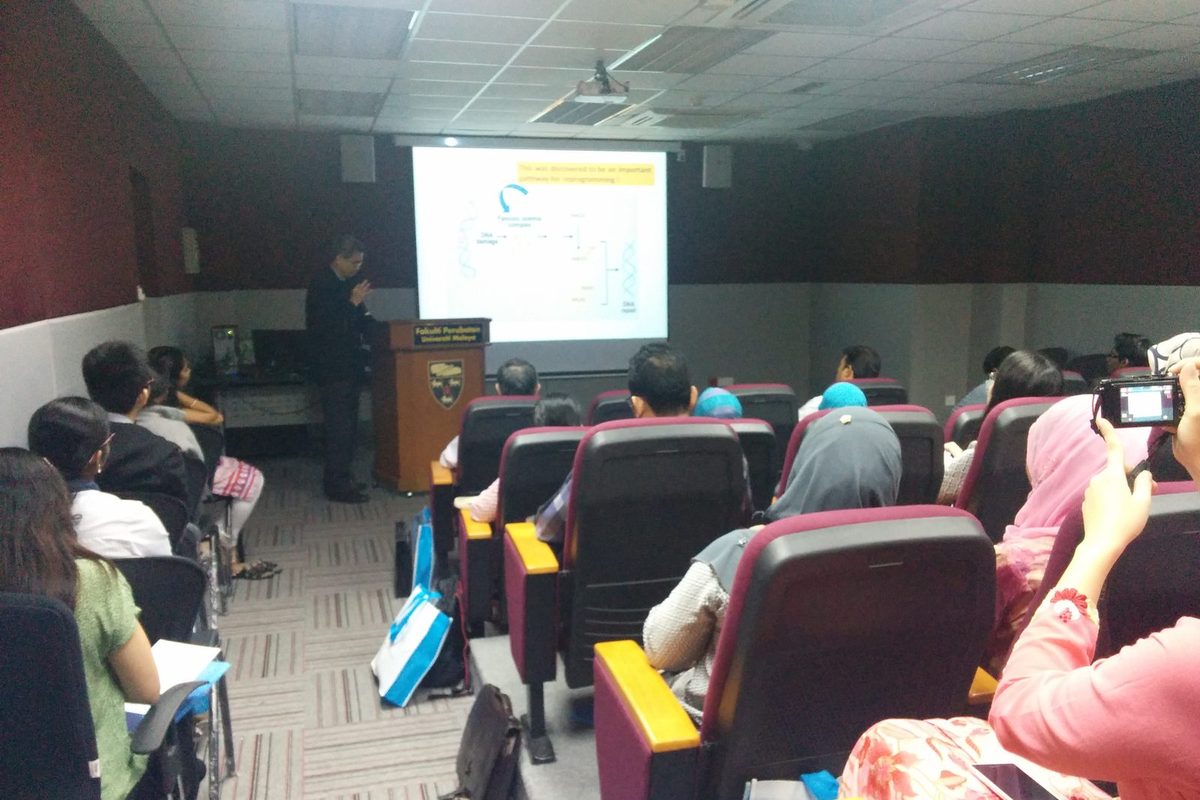 A speaker at a lectern presents a slide to a seated audience in a lecture hall