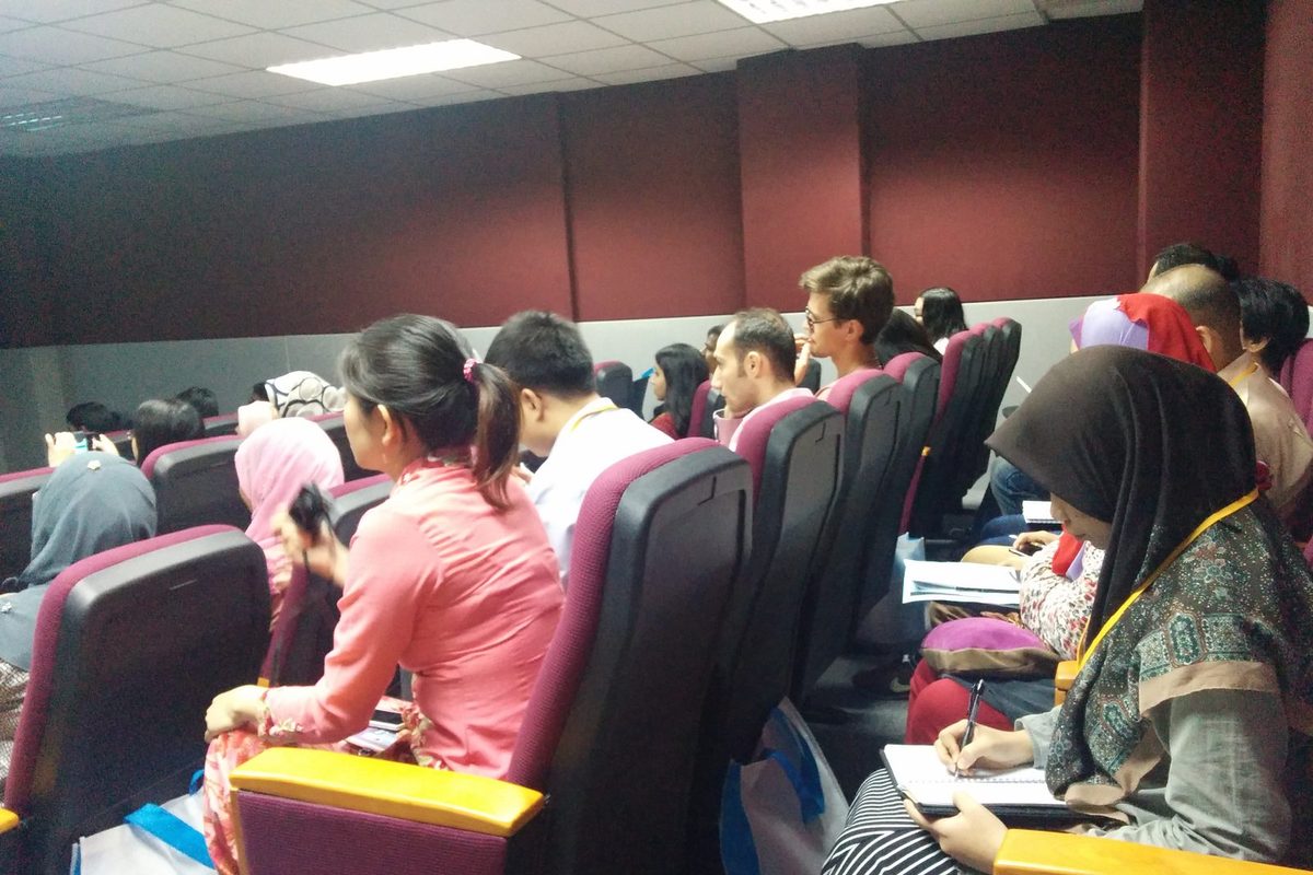 Seated participants in a lecture hall listen and take notes