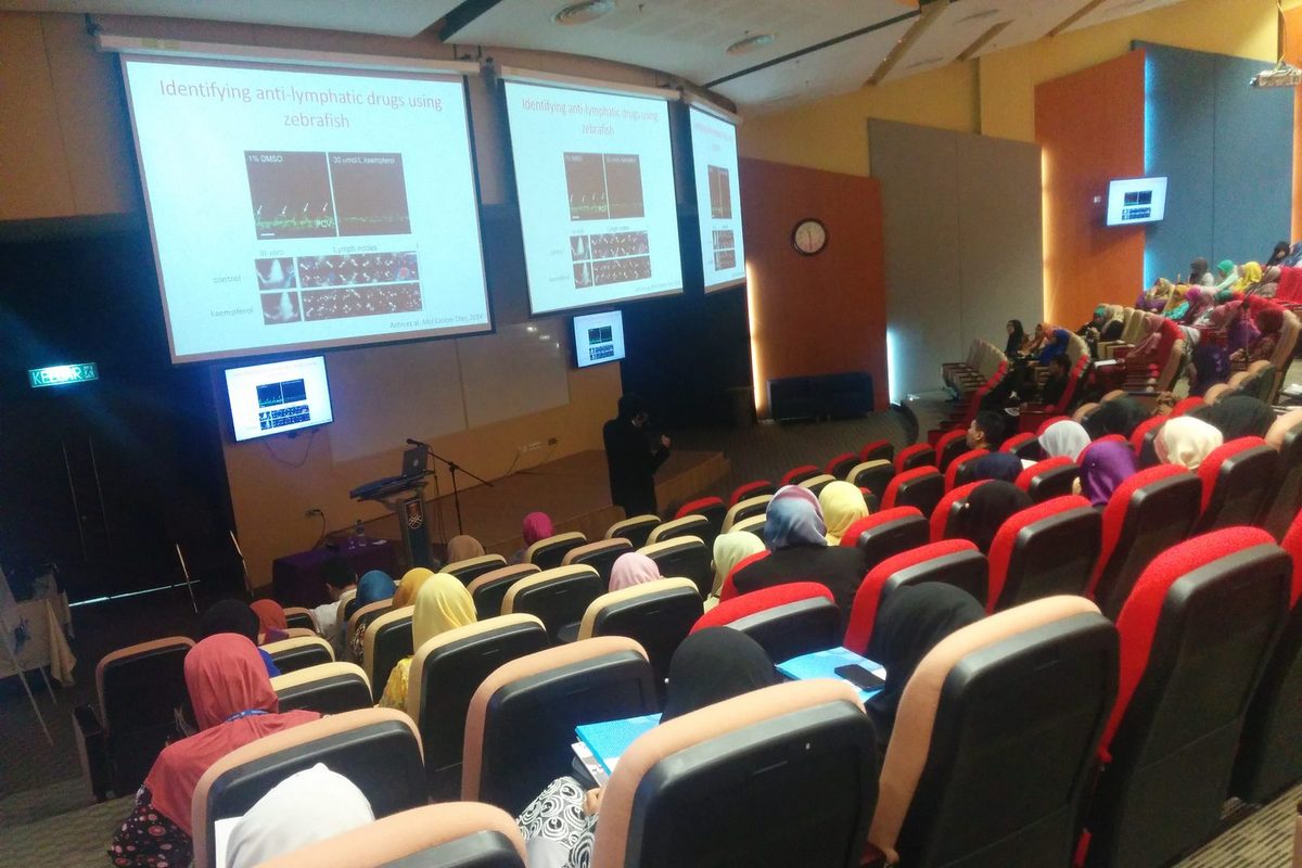 Audience in the auditorium watching a talk, with slides on identifying anti-lymphatic drugs using zebrafish.