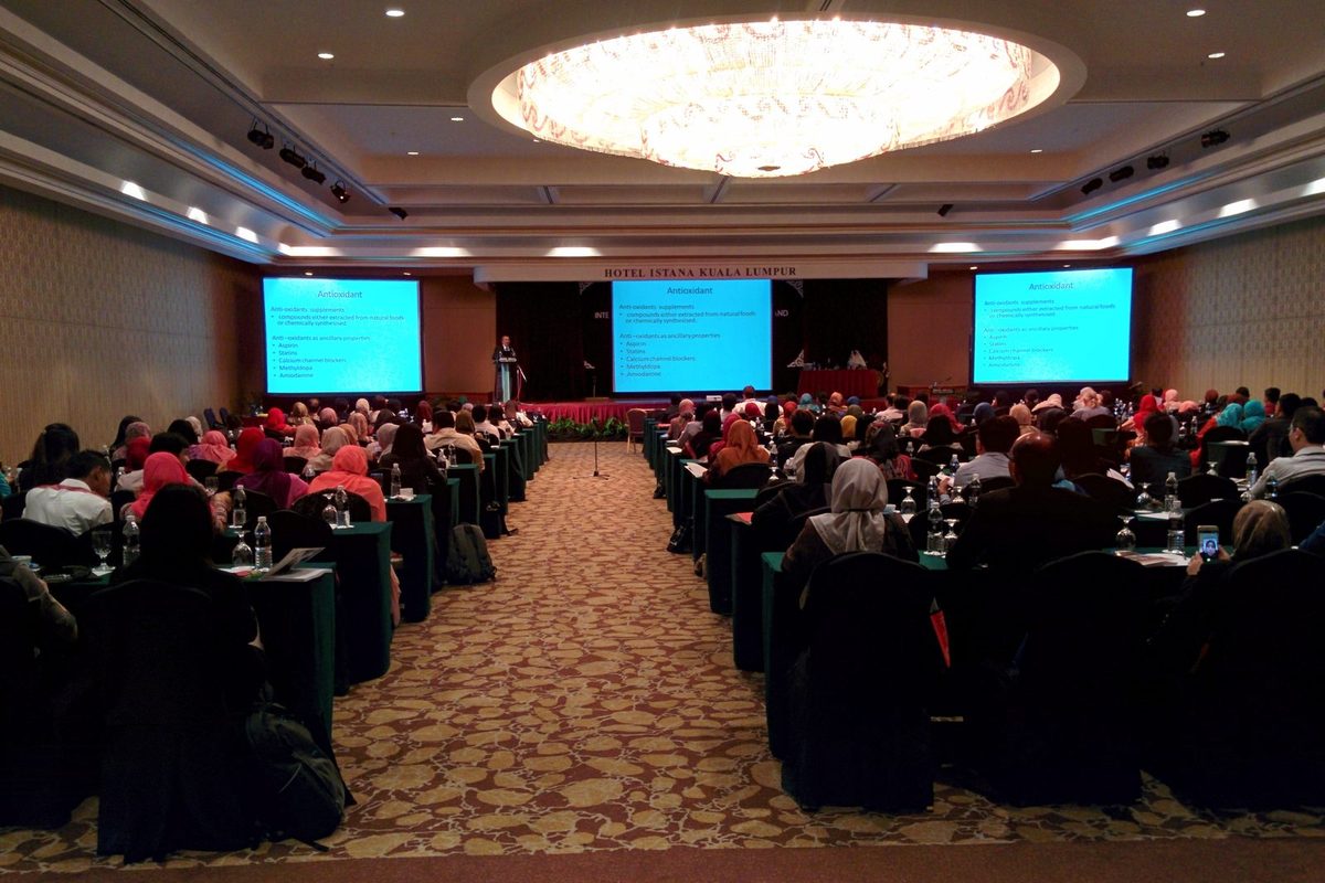 Full conference hall with delegates at long tables facing a speaker and three screens showing a slide on antioxidants