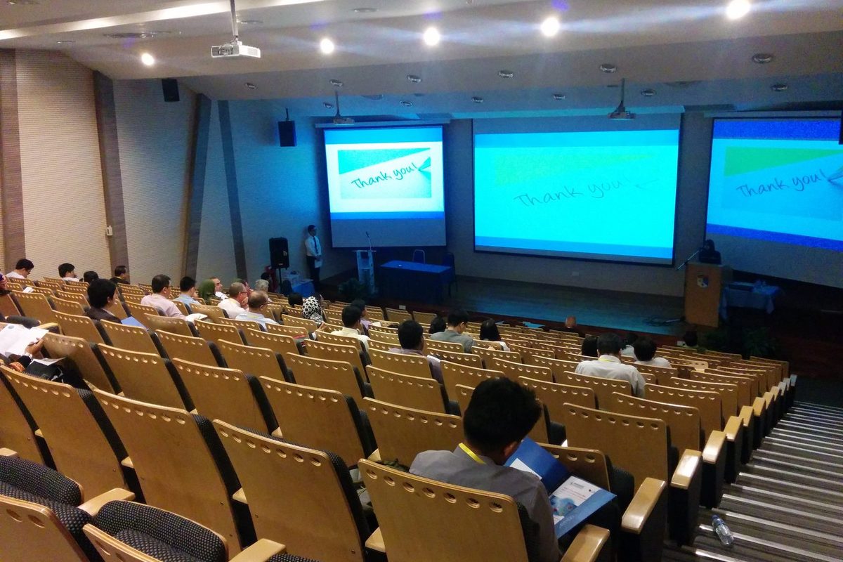 Delegates seated in a tiered lecture hall, facing three large screens