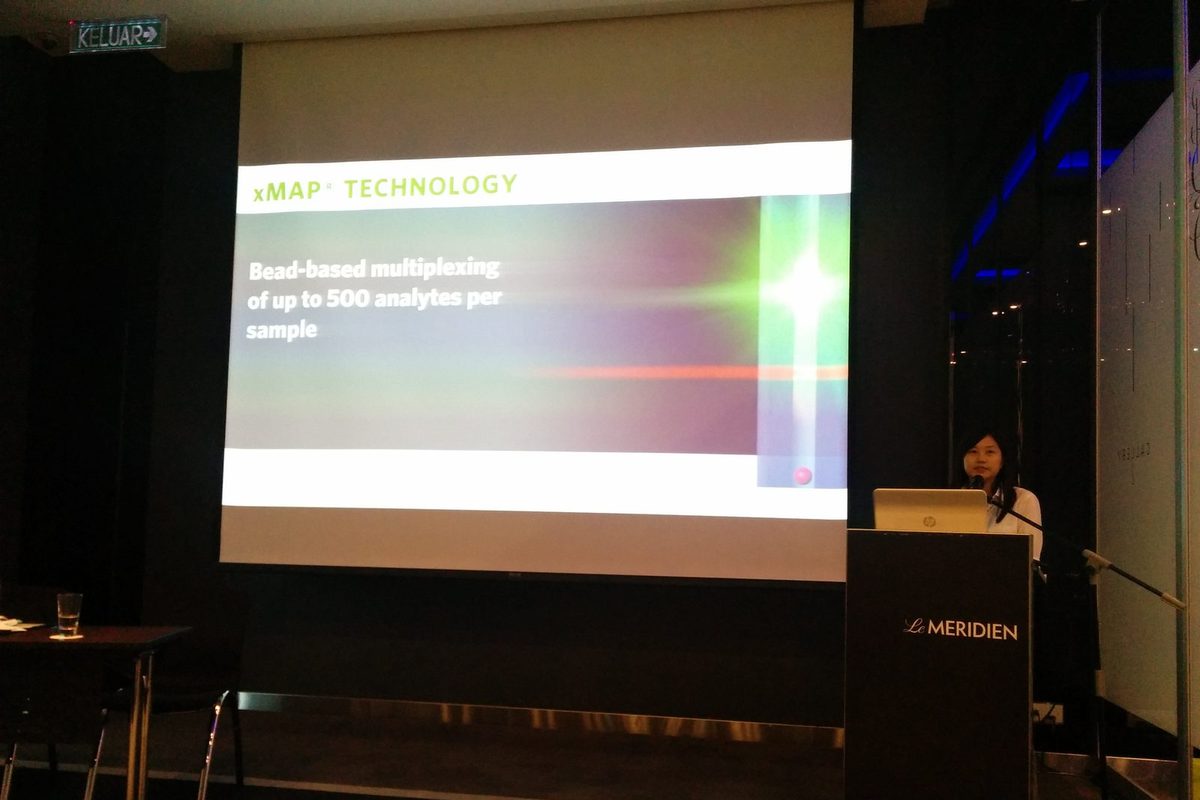 A speaker at the lectern beside a slide titled xMAP Technology, bead-based multiplexing