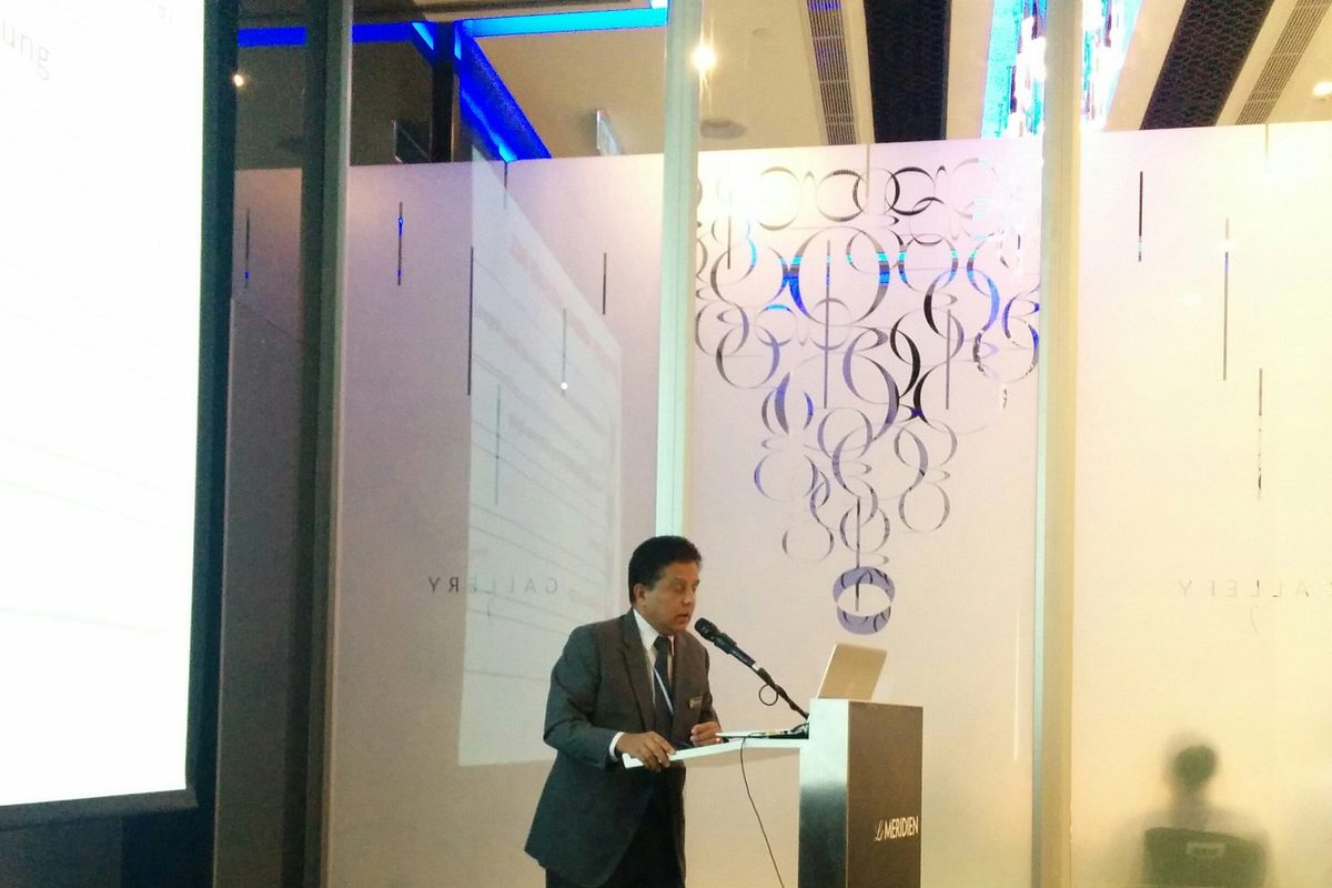 A man in a grey suit speaks at the lectern beside the projection screen