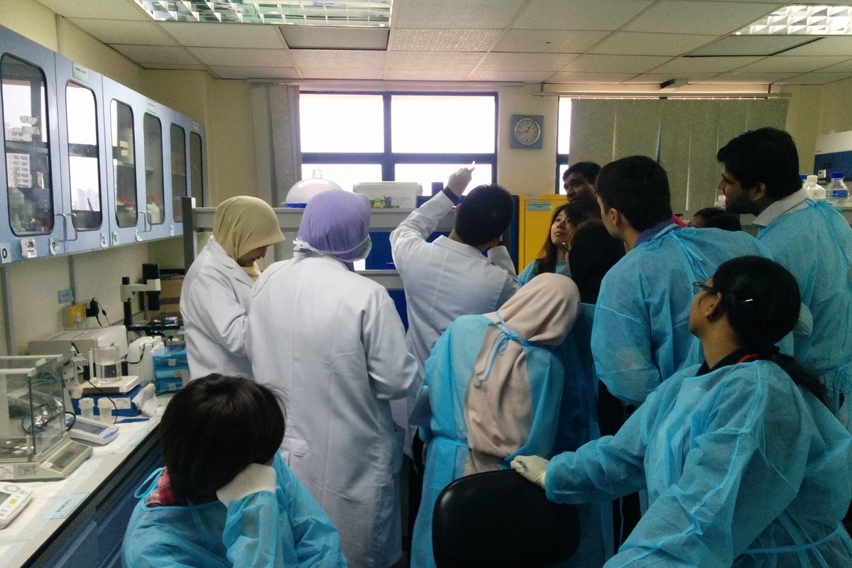 Participants in blue gowns and trainers in white coats around a laboratory bench