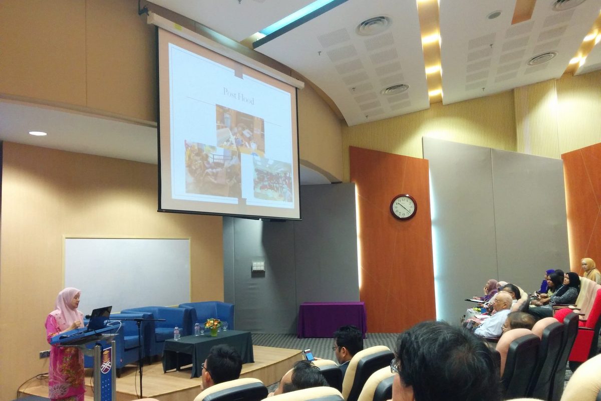 A speaker at the lectern presents a slide titled Post Flood to an audience in a university auditorium