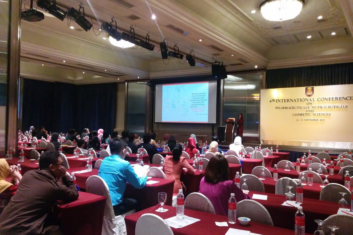 Delegates seated at rows of tables in a hotel ballroom, facing a screen and the conference banner