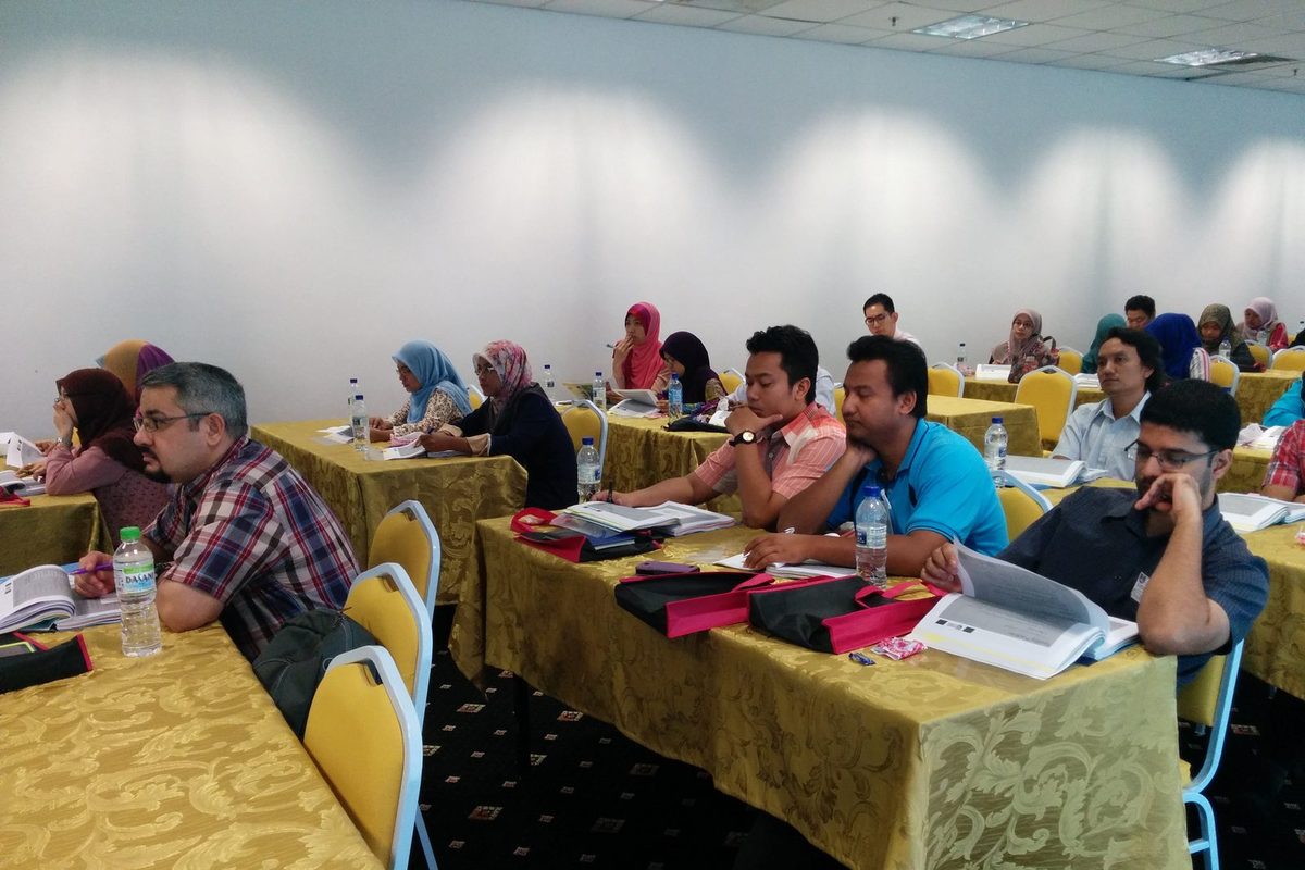 Participants seated at long tables follow a talk with printed notes in front of them