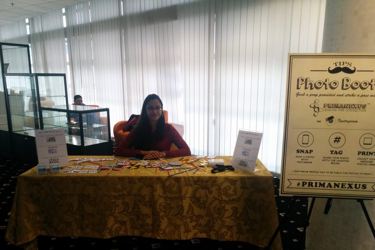 A woman seated at the Prima Nexus photo booth table with props laid out, beside a sign reading snap, tag, print