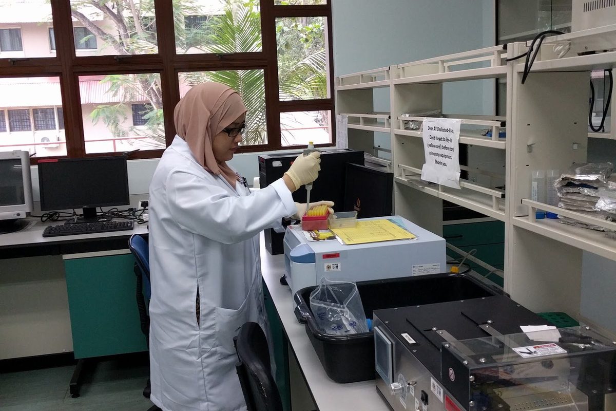 A woman in a lab coat and gloves pipettes at a bench beside laboratory instruments