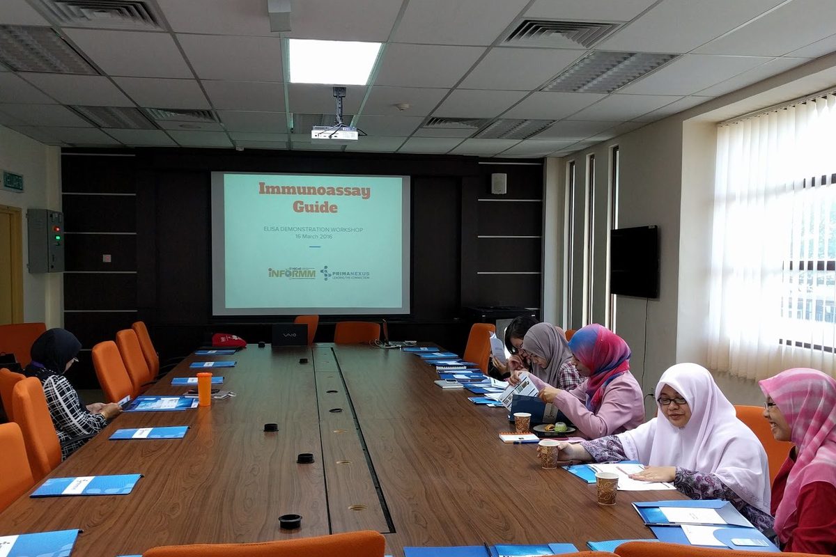 Participants at a long table read their handouts, with the Immunoassay Guide title slide on the screen