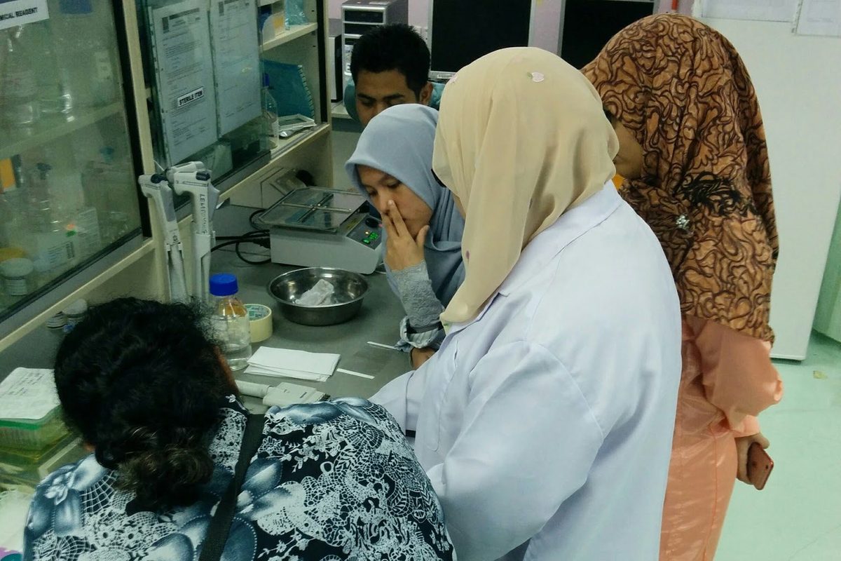 A trainer in a white lab coat works at a laboratory bench while workshop participants look on