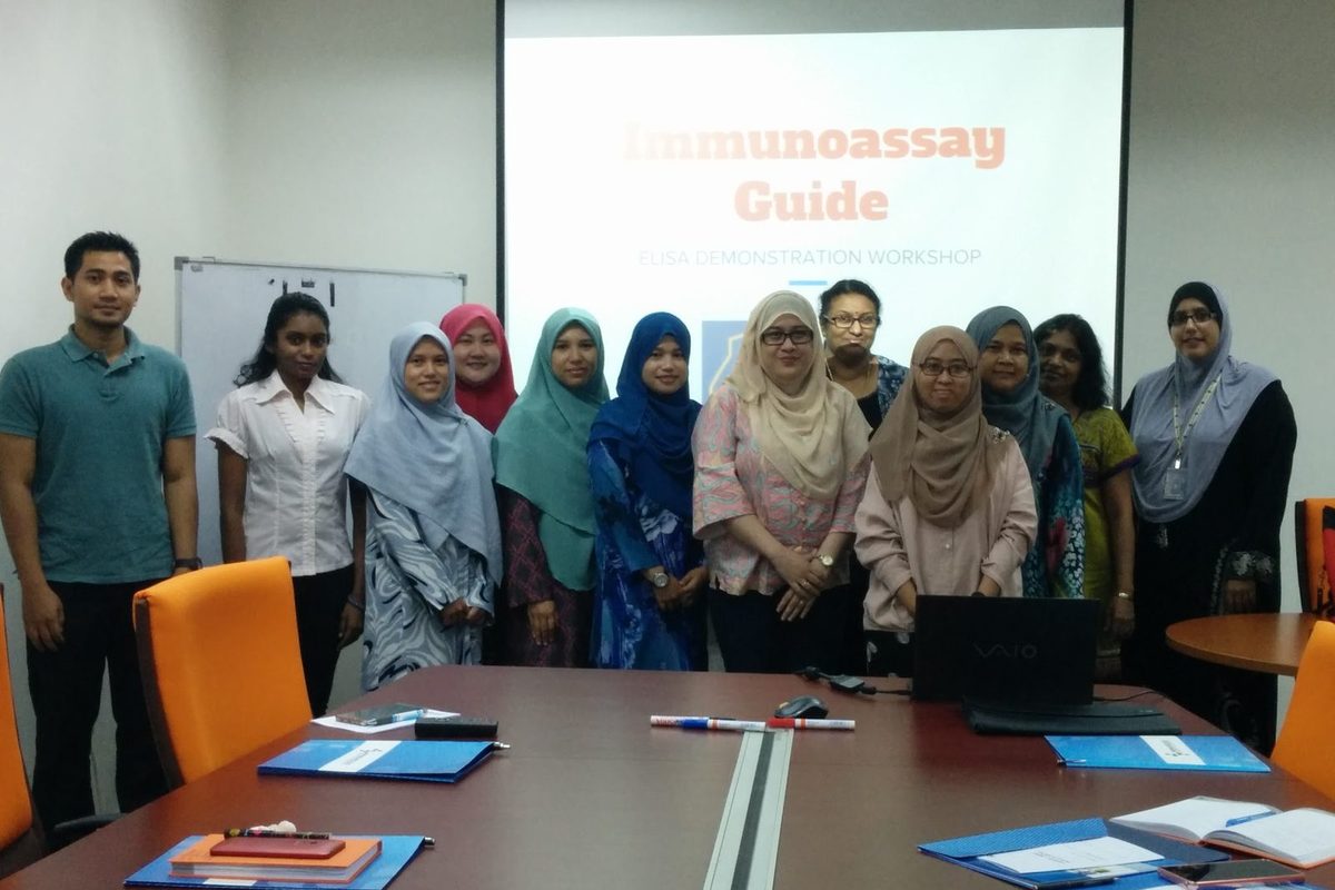 Workshop participants stand together in a meeting room in front of a slide titled Immunoassay Guide
