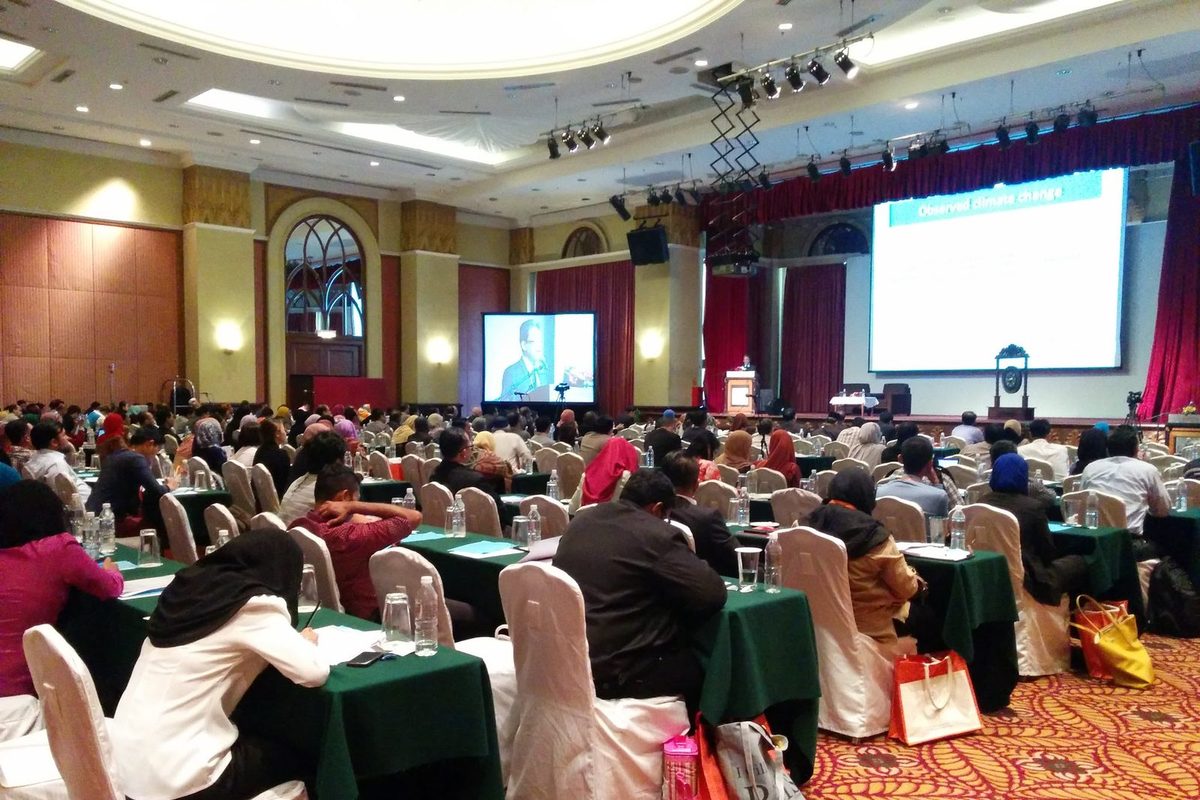 Delegates seated at long tables in a hotel hall, facing a speaker at the lectern and two large screens