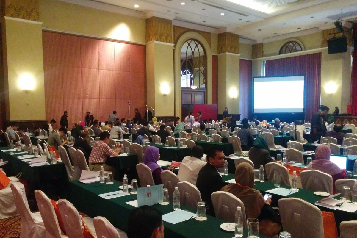 A full conference hall with delegates at long tables facing a projection screen