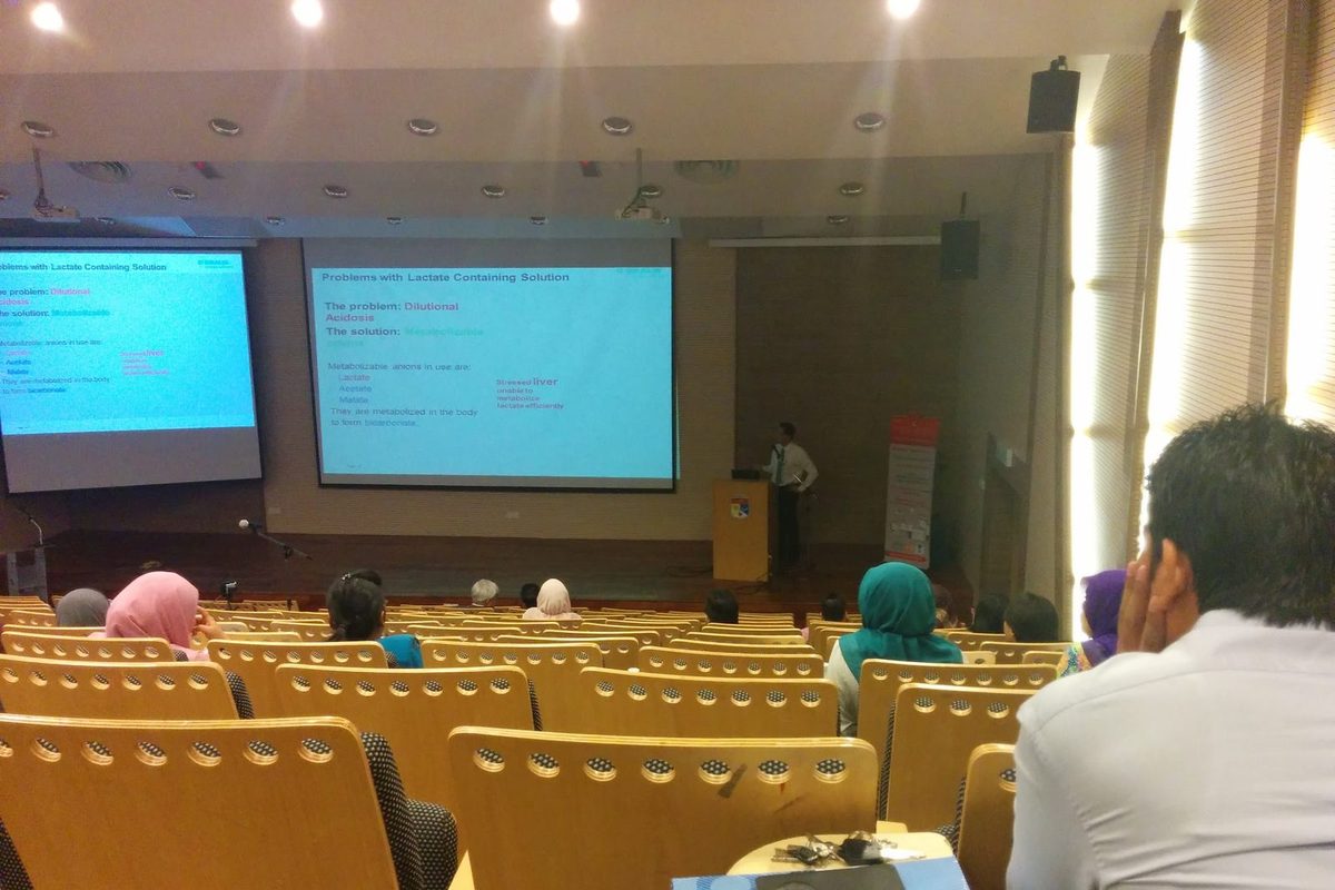 Audience in a lecture hall watching a talk, with slides on two screens and a speaker at the lectern