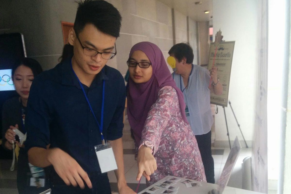 Two people at the booth table looking at printed photos and props, with the Prima Nexus backdrop behind
