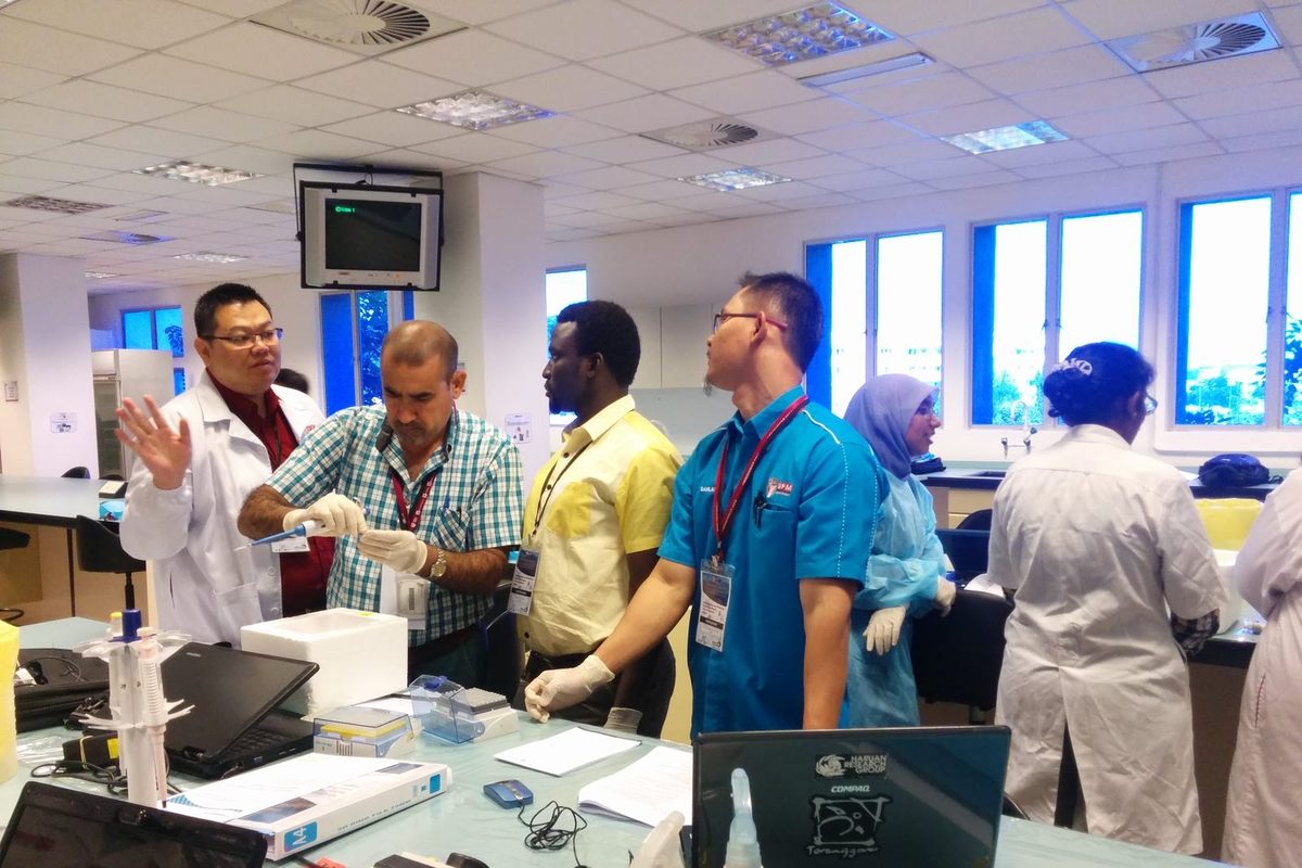 A man in a lab coat explains a step to a group standing at a laboratory bench