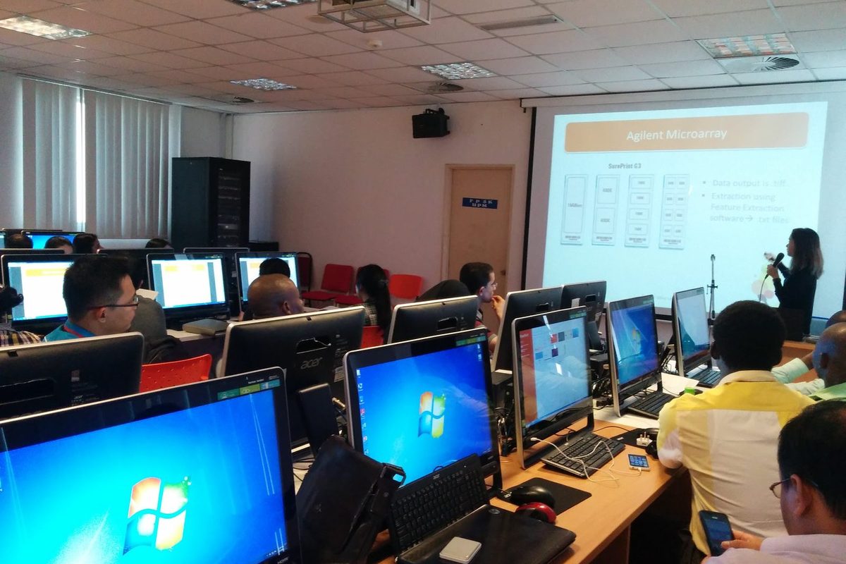 Participants seated at computers in a computer room, with a presenter and a slide at the front