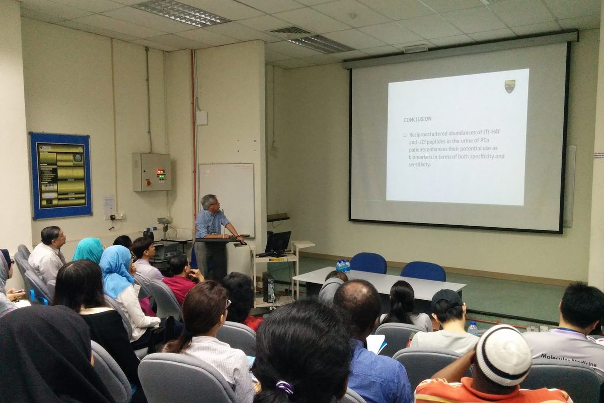 A speaker at the lectern presents a slide to a seated audience in a lecture theatre