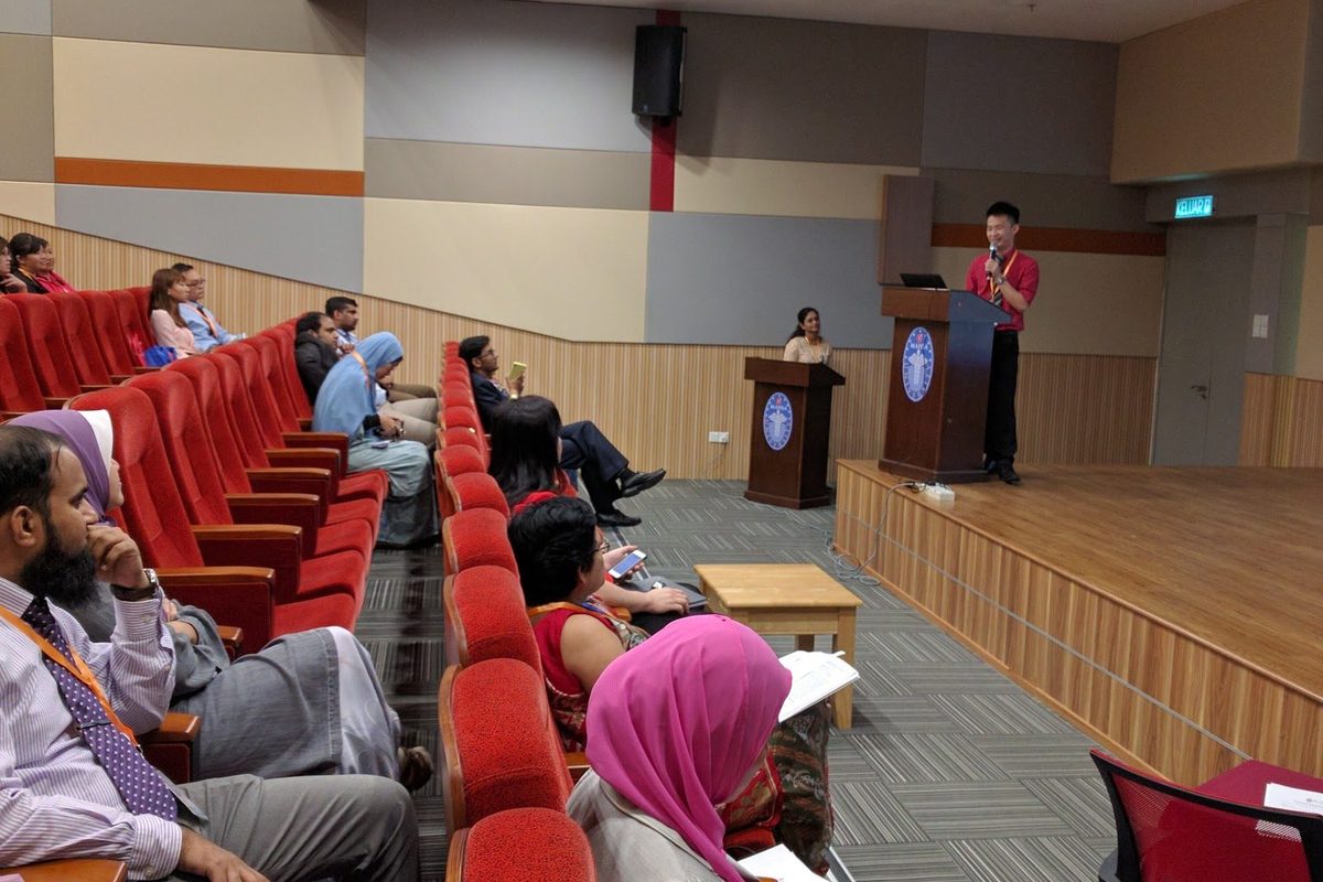 A speaker at the lectern on stage while delegates listen from the red seats of the auditorium