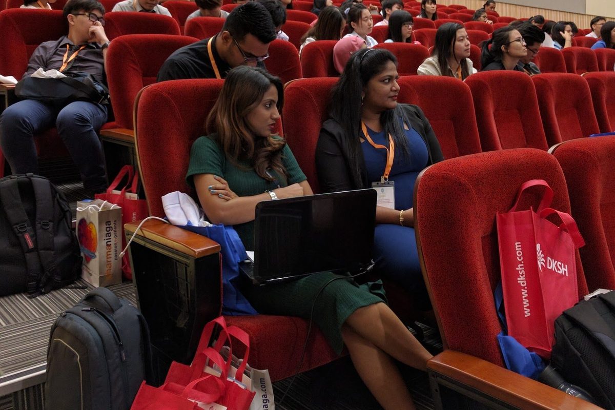 Delegates seated in rows of red seats in the auditorium, some with laptops.