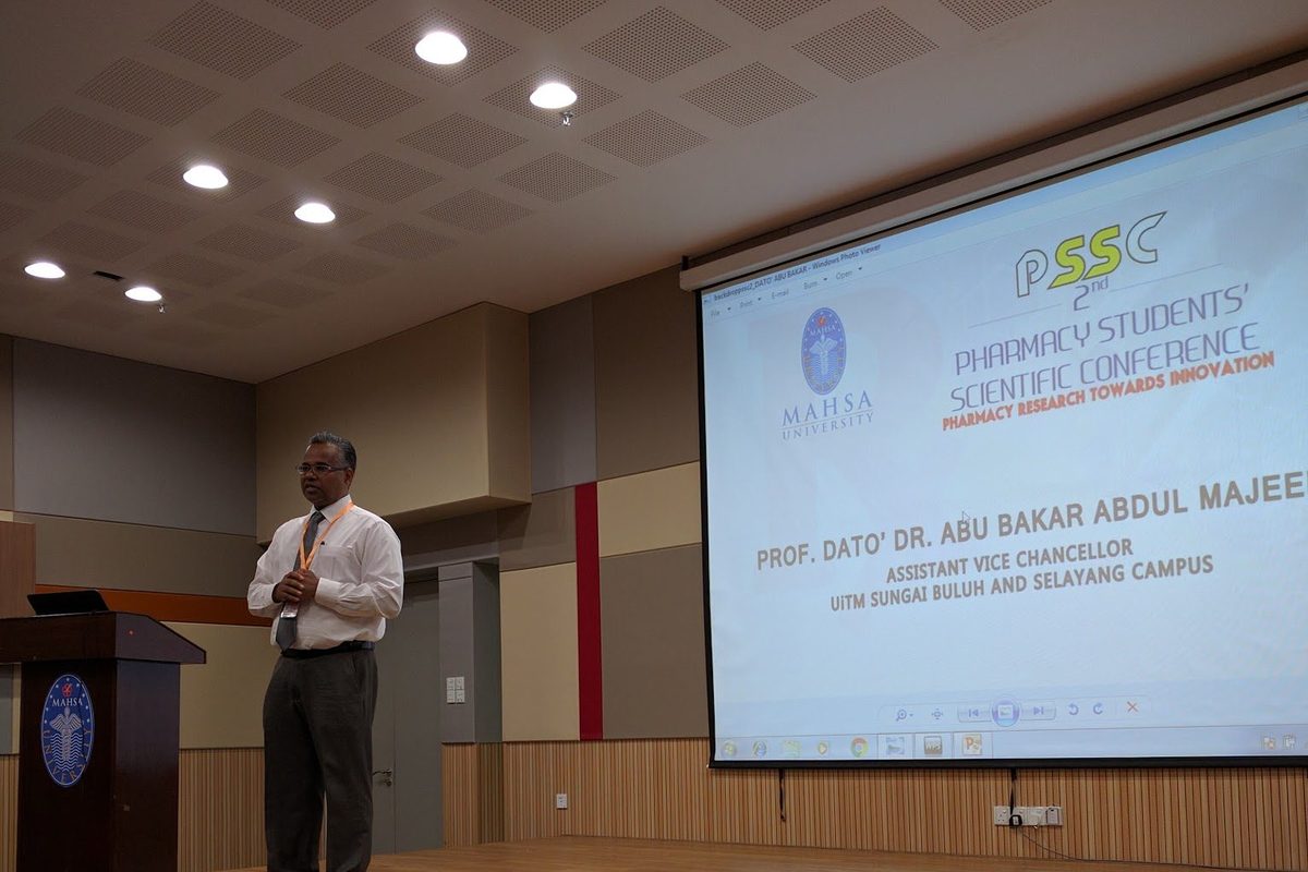 A speaker on stage addresses the audience, with the conference slide on the screen behind him.