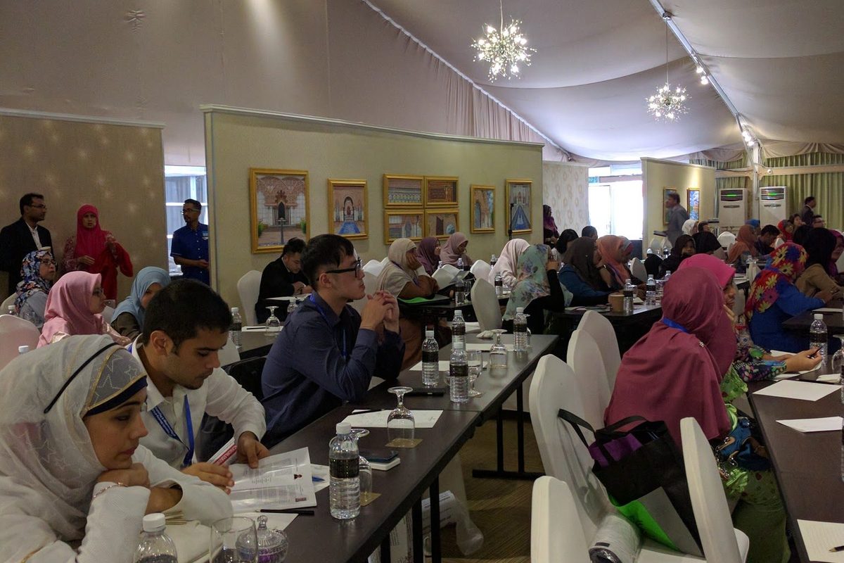 Delegates seated at long tables under a draped ceiling with pendant lights during a session