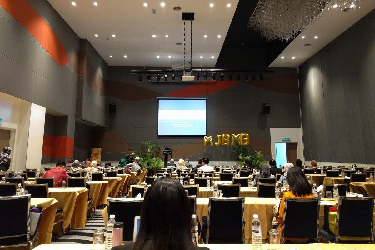 Delegates seated at long tables in the conference hall, facing a screen and a stage with MSBMB balloon letters