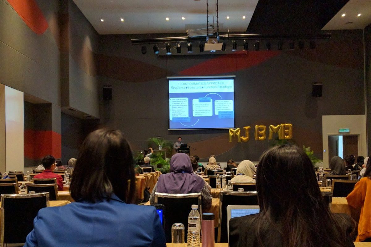 Delegates seen from behind in the hall, with a bioinformatics slide on the screen and a speaker at the lectern