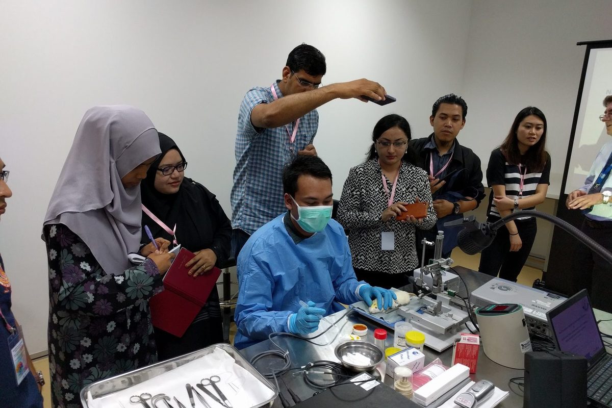 Participants watch a gowned demonstrator at a stereotaxic frame, with surgical instruments laid out on the bench