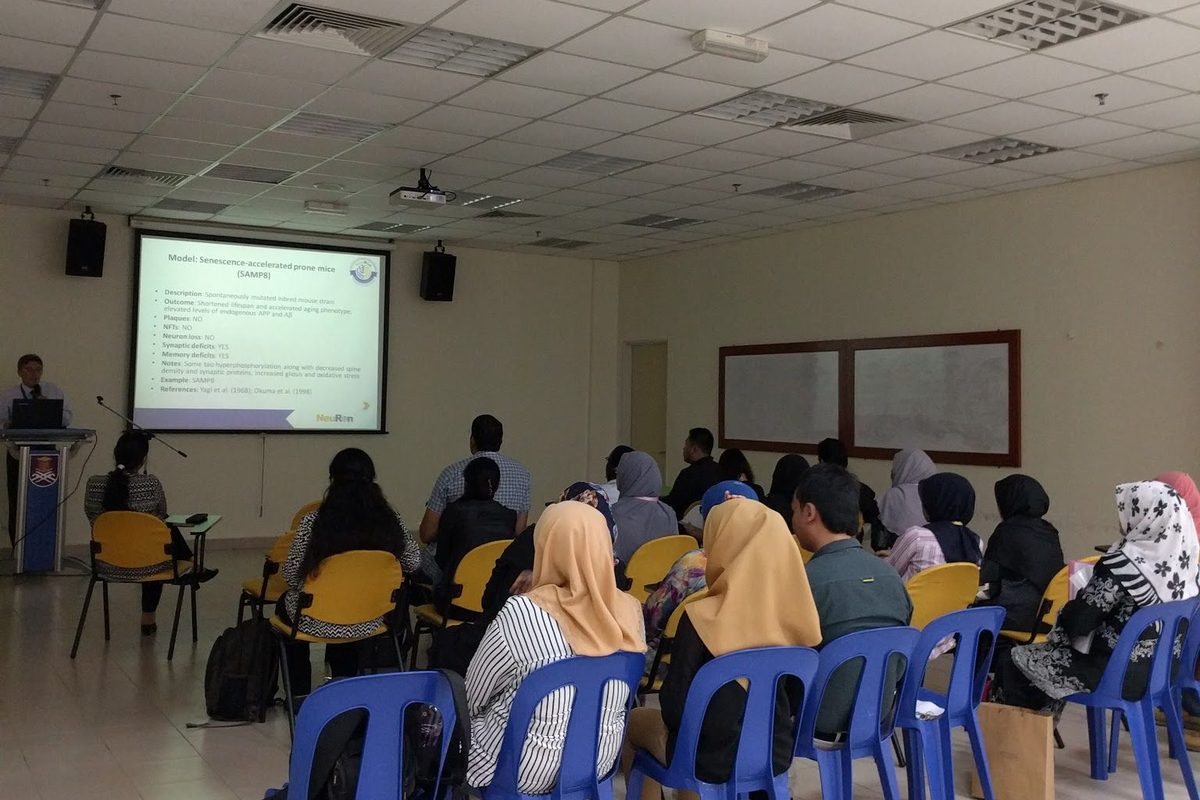 Participants seated in a seminar room listen to a speaker at the lectern, with a slide on mouse models