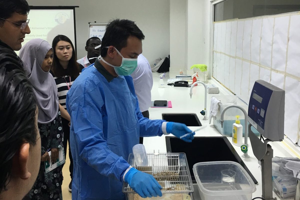 A demonstrator in a blue gown handles a clear rat cage beside a laboratory balance as participants look on