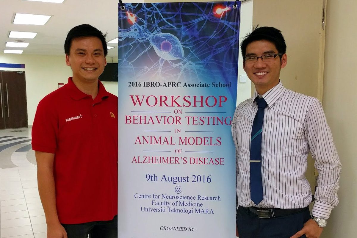 Two men stand beside a tall banner for the workshop on behaviour testing in animal models of Alzheimer's disease