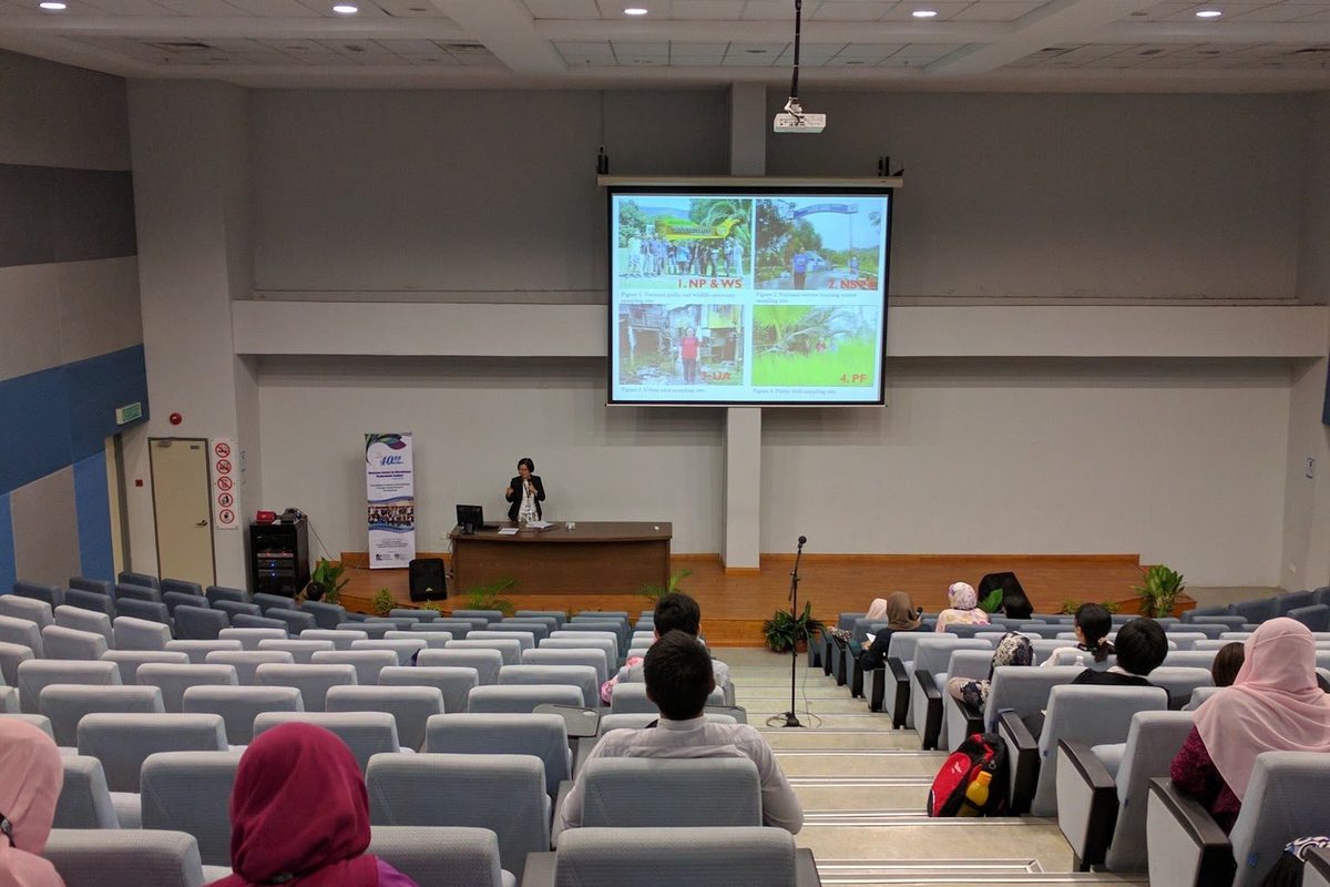 Lecture hall with delegates seated while a presenter speaks from the stage below a projected slide