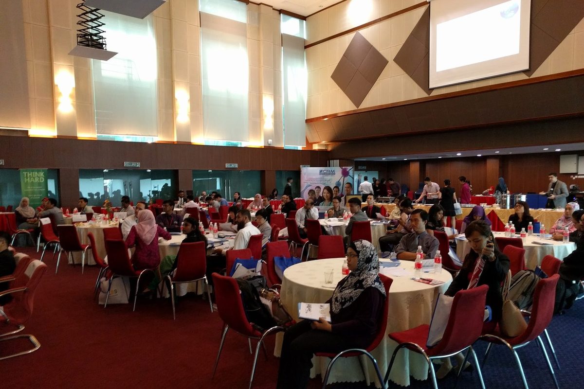Participants seated at round tables in a large ballroom, facing a projection screen