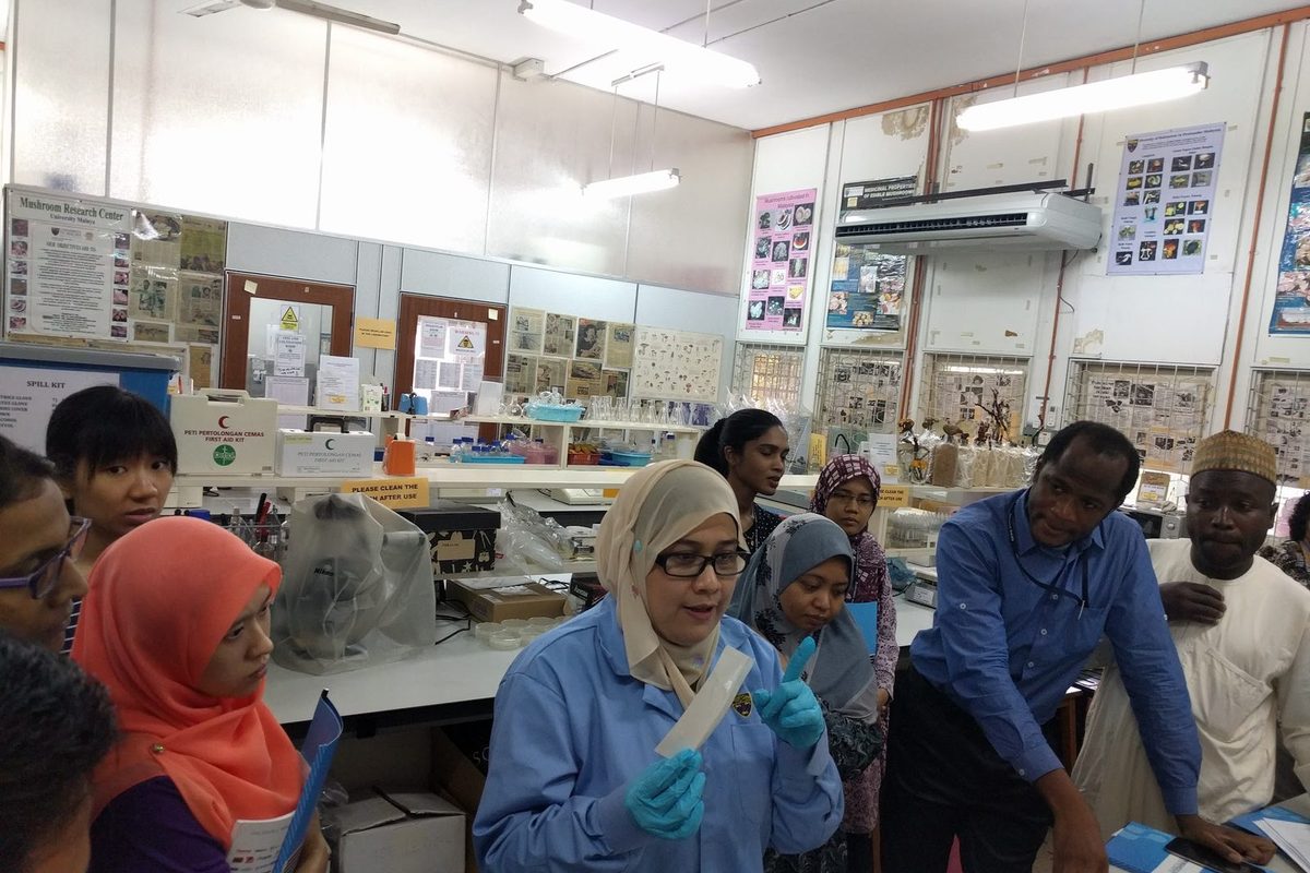 A demonstrator in a blue lab coat and gloves holds up an assay item and explains it to participants at a lab bench