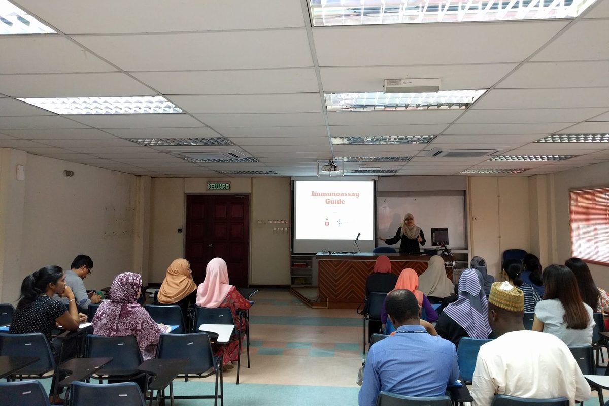 A presenter stands at the lectern beside a projected slide while participants listen in the seminar room.