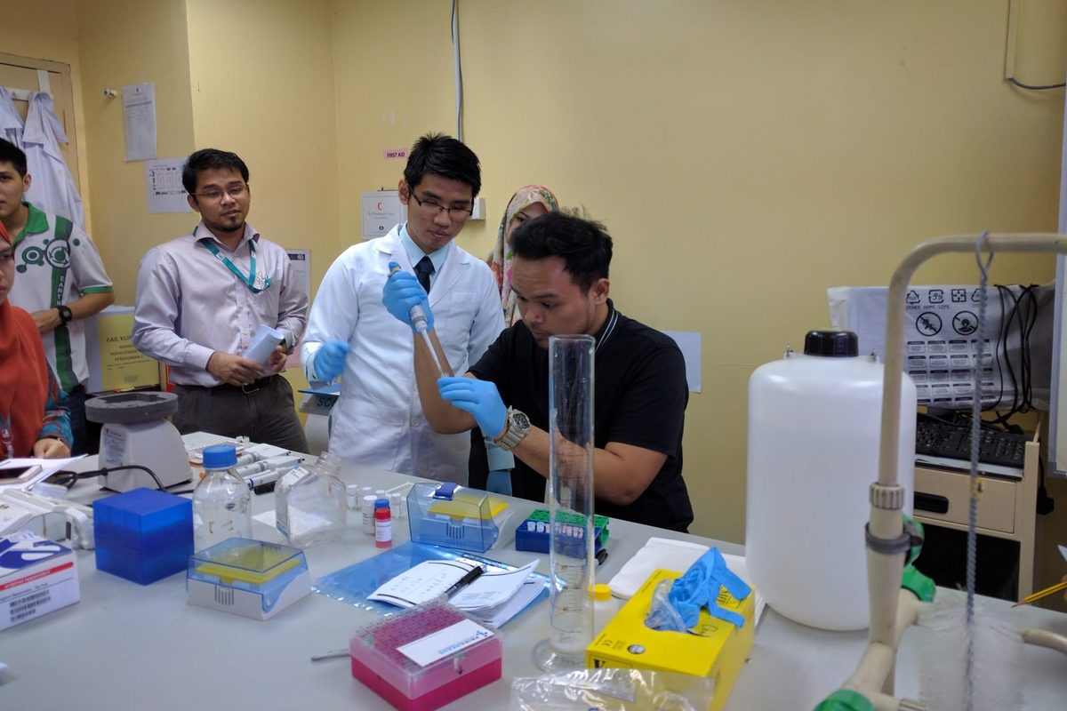 A participant and a trainer in a lab coat work with pipettes at the bench beside a tall measuring cylinder