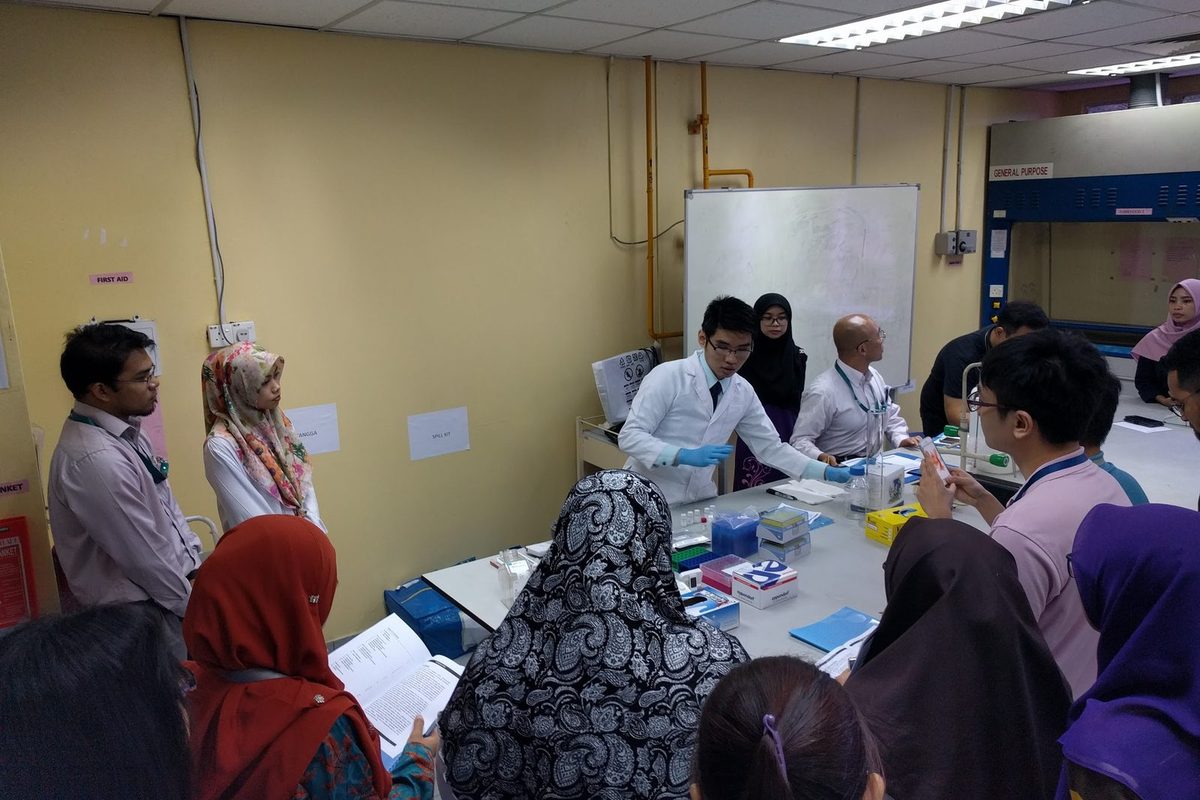 Participants gather around a lab bench to watch a trainer in a lab coat demonstrate an ELISA step