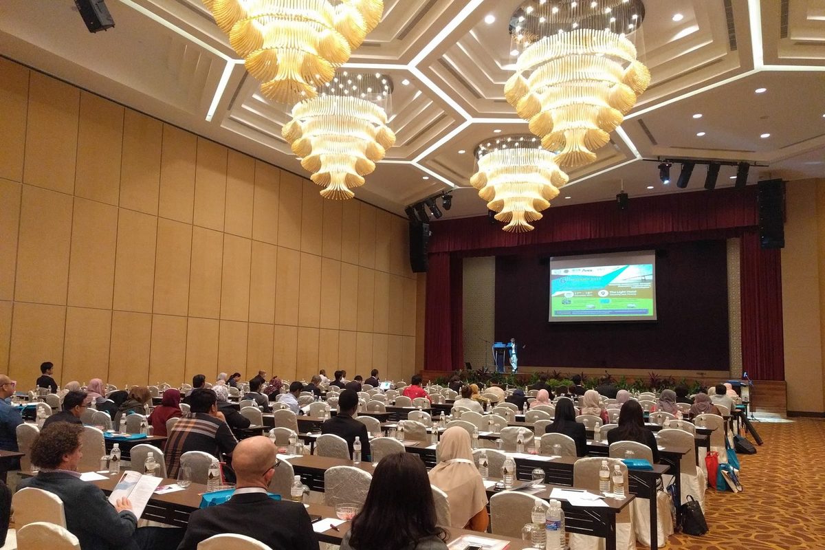 Delegates seated at long tables in a large hotel ballroom, facing the stage and a screen
