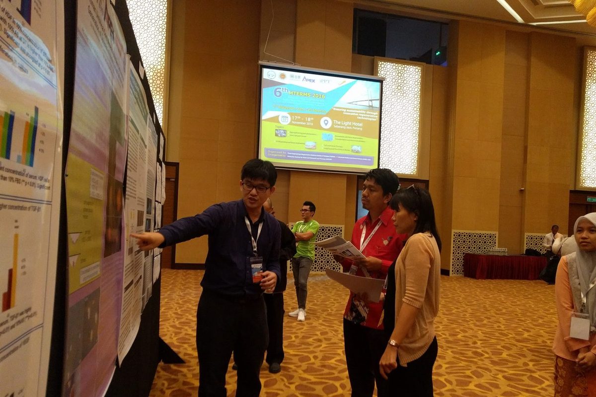 A man points to a research poster as two visitors listen, with the meeting slide on a screen behind