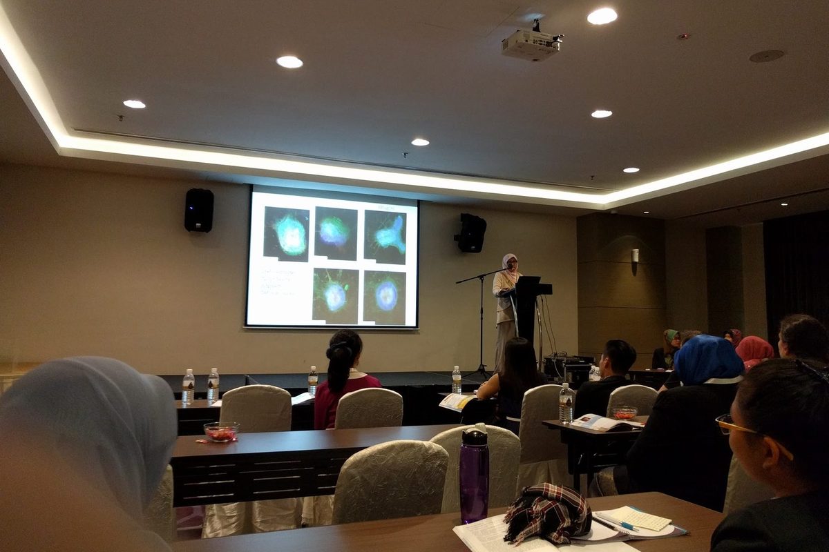 A speaker at a lectern presents microscope images of cells to a seated audience in a smaller room
