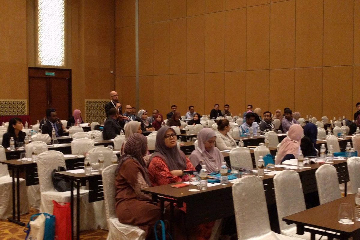 A delegate stands with a microphone to ask a question, with seated delegates around him