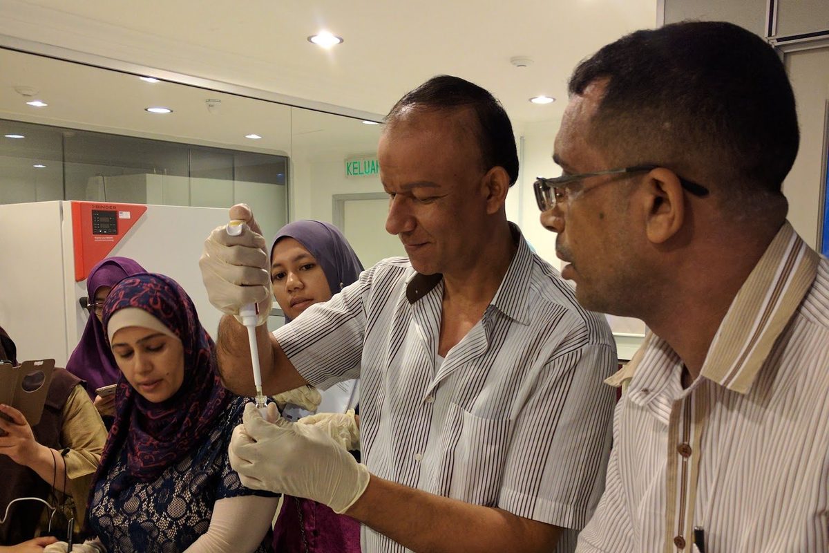A participant wearing gloves uses a pipette at a laboratory bench while another holds the tube and others watch