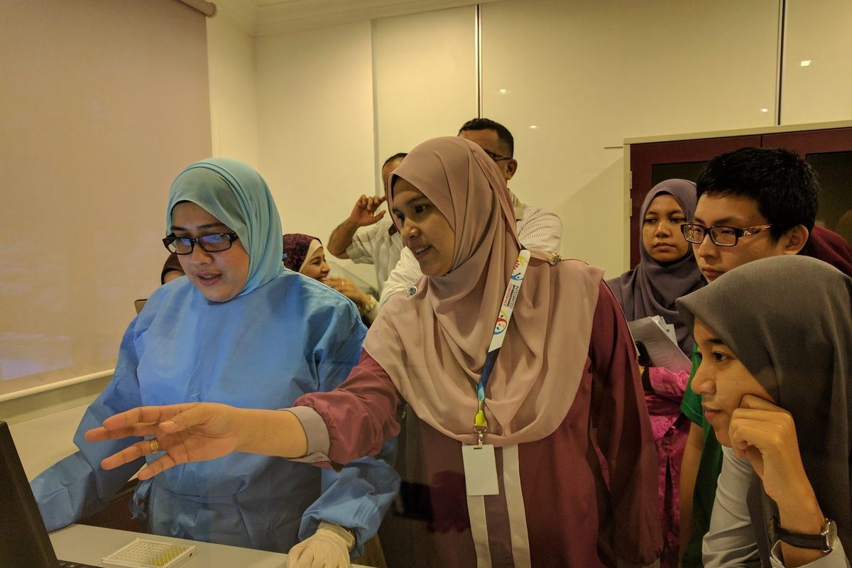 A trainer in a blue lab gown points at a laptop screen next to a 96-well plate as participants look on