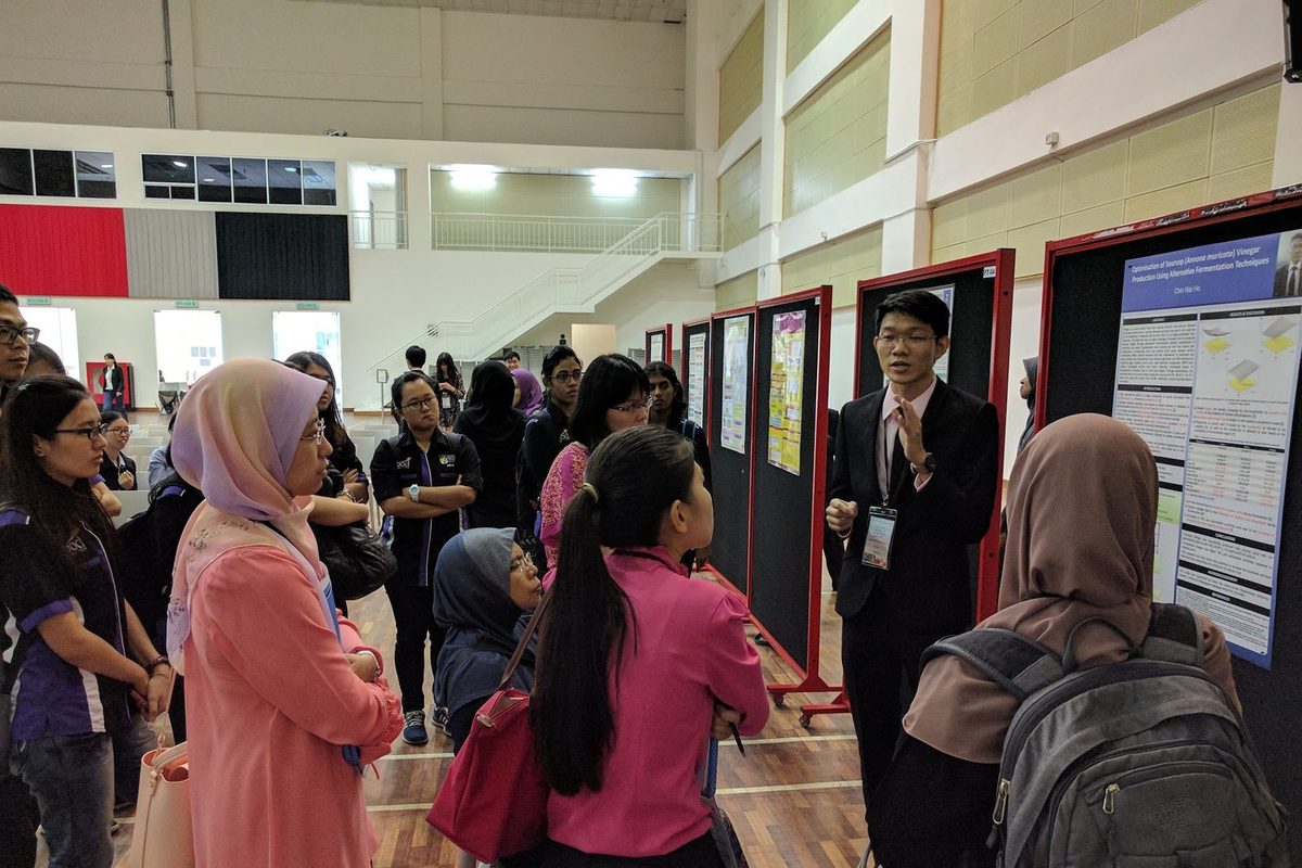 A presenter in a dark suit explains his research poster to a group of visitors in the hall