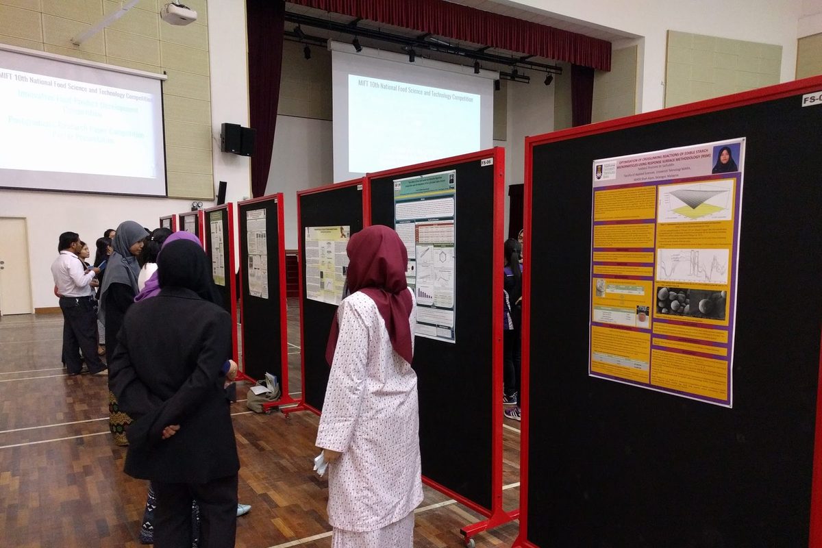 Visitors reading research posters on red-framed boards in a large university hall