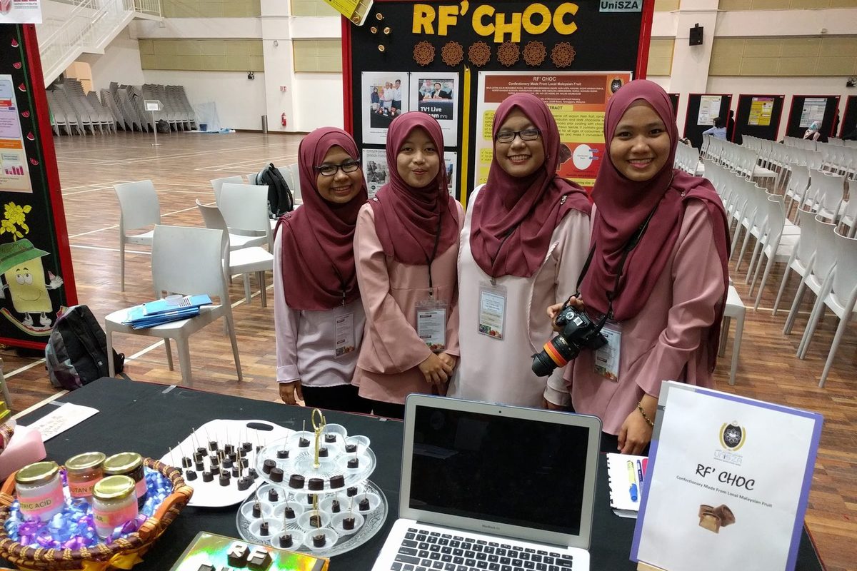 Four students in matching outfits stand behind their chocolate product booth, with a laptop and samples